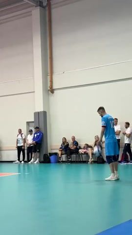 A male volleyball player wearing a light blue jersey with the number 10 on the back and matching light blue shorts, is in the process of serving a volleyball. He is holding the ball in his left hand and his right arm is extended backwards, preparing to strike the ball. The player is on a teal colored volleyball court with a red boundary line visible. In the background, several other individuals are standing and sitting, observing the game. One person is standing on a red referee stand near the net. The environment appears to be an indoor gymnasium with a high ceiling and bright overhead lighting.