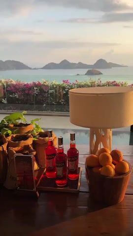 The video shows a table with several objects on it, set against a backdrop of a scenic view of islands and the ocean. On the table, there are three bottles of Aperol, a wooden bowl filled with oranges, a lamp with a beige shade, and some potted plants. In the background, there is a body of water with islands in the distance. The sky is cloudy, and the sun appears to be setting, casting a warm glow over the scene. The overall ambiance is serene and relaxed, suggesting a vacation or a leisurely moment.