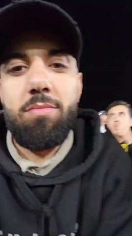Here's a concise description of the video:

The video begins with a close-up of a man with a beard and a black baseball cap wearing a black Adidas hoodie, smiling at the camera. The camera then pans out to show the man in the stands of a stadium, likely at a sporting event, possibly soccer. The stadium is full of people, and there are players on the field. Several other people are standing or sitting nearby in the stands. The stadium lights are on, illuminating the field.