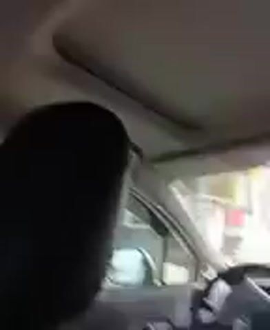 The video shows a young child with pink and brown hair sitting in the back seat of a car. The child is looking at the camera and smiling. The interior of the car is visible, including the headrests of the seats and the ceiling. The child is wearing a light-colored shirt. Outside the car window, buildings and another vehicle can be seen. The child smiles broadly and laughs.