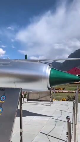 The video pans across a memorial with a silver fighter jet on display. A plaque lists battle and theater honors. In the background, mountains are visible under a blue sky with clouds. An Indian flag flies on a pole. The jet has Indian Air Force roundels.