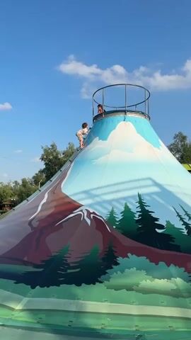 A vertical, low-angle video shows two children climbing an inflatable volcano-shaped slide under a bright blue sky with scattered clouds. The slide is decorated with a painted landscape featuring a snow-capped mountain and pine trees. One child, wearing a light-colored shirt and shorts, is actively climbing the side of the volcano using a rope ladder. Another child, visible at the top of the slide, is looking down and wearing a pink shirt. The ground area is out of focus.