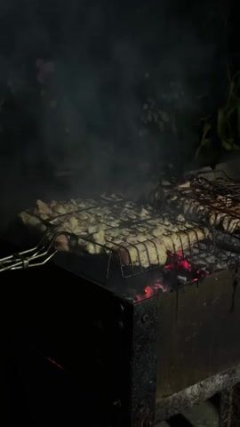 The video shows meat grilling on a barbecue at night. The barbecue is a dark metal box with glowing red coals visible underneath the grill grates. Two wire grill baskets are filled with pieces of meat, some of which appear to be chicken. Smoke rises from the meat as it cooks. The background is dark, with some blurry green foliage and hints of flowers visible. The scene suggests an outdoor gathering or meal preparation.