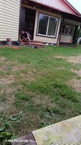 The video shows a man in a dark t-shirt and shorts working outside a small building. He is kneeling on the ground, appearing to be engaged in some sort of repair or construction work. There are various tools and materials scattered around him, including planks of wood, a red bucket, and what looks like a brush. The building has a light-colored exterior and a porch with a red roof. The ground is covered in grass and dirt. The overall scene suggests a casual outdoor work environment.