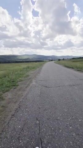 A first-person perspective video shows a person riding a bicycle down a paved road. The road is cracked and weathered. On the left side of the road is a field of green grass. On the right side of the road, the land slopes upwards, also covered in grass. In the distance, there are rolling hills and a town or cluster of buildings. The sky is partly cloudy with white clouds against a blue sky. The handlebars of the bicycle are visible at the bottom of the frame.