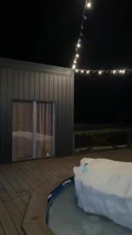 At night, a wood deck illuminated by a string of lights reveals a cabin-like structure with a sliding glass door. To the right, a hot tub is partially covered with snow. The surrounding area beyond the deck is dark, but hints of greenery and a distant structure are visible.