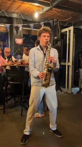 A young man with curly blonde hair stands in the center of the frame playing a tenor saxophone. He is wearing a light blue button-down shirt and light beige pants. He is looking straight ahead. To his left, there are several people sitting at a high-top table, including a man with glasses and a woman with red hair. Behind the saxophonist, there is a woman with dark hair. To the right of the saxophonist, a person wearing a patterned shirt walks by, partially obscuring the view. The environment appears to be an indoor venue with a wooden ceiling and string lights. There are also speakers and a doorway visible in the background.