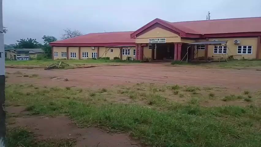 The video shows a wide shot of a building labeled "FACULTY OF FOOD AND CONSUMER SCIENCES". The building is one story and has a red roof and yellow walls. There are several windows and air conditioning units on the exterior. The building is surrounded by a dirt area with patchy grass. The sky is overcast.