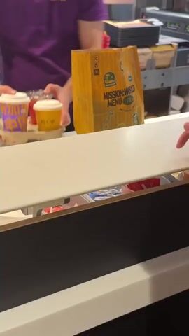 The video shows a close-up of a fast-food restaurant counter. In the foreground, there is a white counter surface. Behind it, on a tray, are two McCafe cups and a drink in a clear plastic cup. The McCafe cups have white lids and one is purple with Chinese characters, while the other is yellow with the McCafe logo. The clear cup appears to contain a red beverage. To the right of the cups is a large, yellow McDonald's paper bag with "MISSION-WORLD MENU" printed on it. A portion of a person wearing a purple shirt is visible on the left side of the frame, their arm and hand are reaching towards the tray of drinks. In the background, there are stacks of items, possibly food packaging, and a red container. The environment appears to be a busy service area of a fast-food establishment.