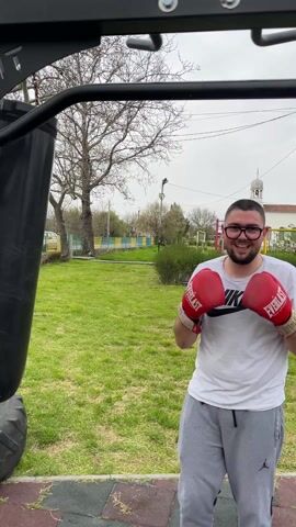 A young man wearing glasses, a white Nike T-shirt, and gray sweatpants is standing next to a black punching bag outdoors. He is wearing red Everlast boxing gloves and smiles at the camera. The background shows a grassy area, trees, and a building with a white dome. The camera zooms in on the man's face and then shows him in a boxing stance. The day appears overcast.