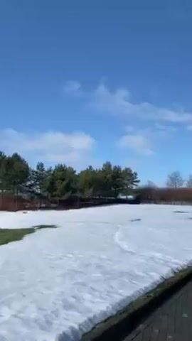 The video shows a winter scene. A thin layer of ice covers a body of water. Patches of snow are visible on the ground. The sky is blue with scattered white clouds. Trees line the horizon in the background. Some trees are bare, while others are evergreen. The sun is partially obscured by the clouds. The scene is still and quiet.