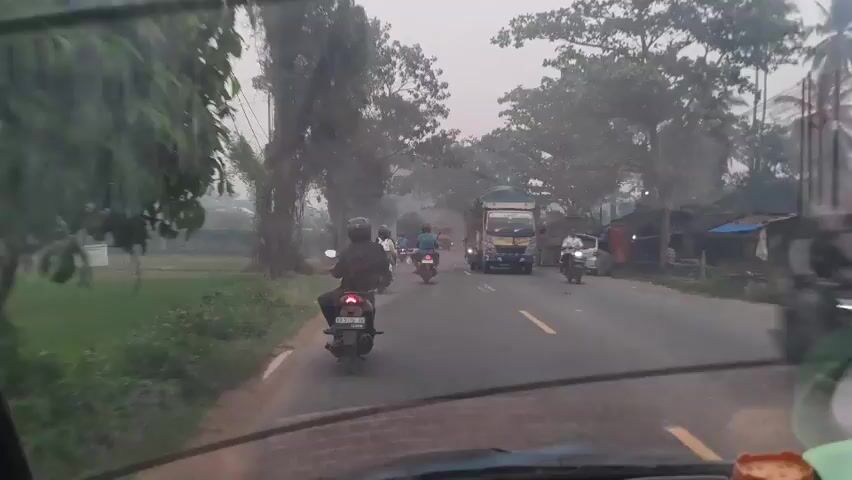 The video shows a view from inside a vehicle, looking through the windshield. A road is visible ahead, lined with trees and greenery on both sides. Several motorcycles and a truck are traveling on the road. Some of the motorcycles are moving away from the camera, while others are approaching or moving parallel. The truck is a large, yellow vehicle with a green tarp covering its cargo. There appears to be some haze or fog in the environment, particularly on the left side of the road where there are fields and what might be a small building. The sky is overcast. The time stamp on the video is 00:00 to 00:05.
