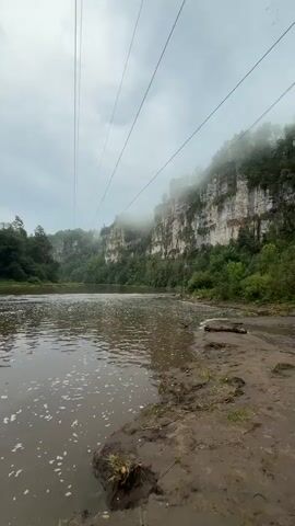 A wide, muddy river flows towards the viewer in the foreground. The water is choppy and has a foamy surface. On the right bank, there is a muddy shore with some scattered leaves and debris. In the background, on the left side of the river, there is a dense forest with tall green trees. On the right side of the river, imposing, steep, rocky cliffs rise up, partially obscured by mist and fog. The sky is overcast and gray. Several thin, dark power lines stretch diagonally across the upper portion of the frame from left to right.