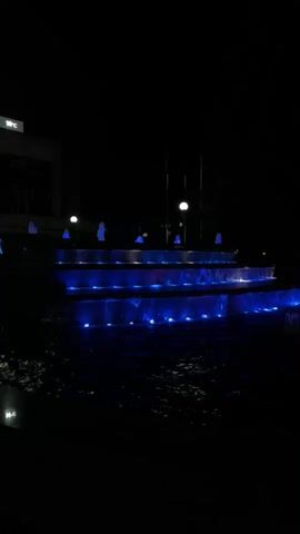 The video shows a tiered fountain at night. The fountain is illuminated with blue lights. In the background, there is a building with some lights on. To the right of the fountain, there is a colorful water fountain. The scene is dark and the water is sparkling.