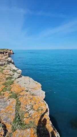 The video shows a beautiful, rocky coastline. The camera is positioned on a cliff, looking out over the turquoise-colored sea. The sky is clear blue with a few wispy clouds. The cliffs are composed of layered rock formations, some covered in orange lichen and green vegetation. The sea appears calm and stretches to the horizon. There are no people visible in the video. The focus of the video is the natural beauty of the coastal landscape.