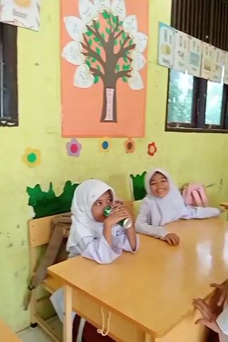 Two young girls wearing white hijabs and uniforms are in a classroom. One girl on the left is drinking from a green can. Another girl stands to her right, also in uniform, and appears to be talking or gesturing. A third girl is seated at a desk further back, reading a book. The classroom walls are yellow and decorated with colorful drawings, including a tree with leaf-shaped cutouts and what appear to be alphabet charts above a window. Desks are made of light-colored wood.
