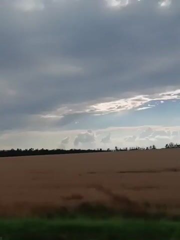 This video captures a drive through a rural landscape under a cloudy sky. The scene is dominated by a vast, golden field, likely agricultural, stretching towards a tree line in the distance. The sky is filled with dramatic, gray clouds, with some hints of sunlight breaking through, suggesting either early morning or late afternoon. There are no people visible in the footage.

The perspective shifts, showing the view from inside a vehicle. We see the dashboard and part of the steering wheel, indicating the drive is in progress. The windshield wipers are visible and appear to have recently been used, suggesting light rain or dew. Outside, the road is paved, and another vehicle can be seen ahead, moving in the same direction. Roadside vegetation, including green grass and some trees, lines the path. A traffic sign is briefly visible on the right side of the road.

The overall atmosphere is serene and perhaps a bit somber due to the overcast sky, but the expanse of the field and the open road evoke a sense of journey and freedom. The location context of Rozovka, Russia, places this scene in a potentially agricultural region of the country, consistent with the visual evidence of farmland. There is no visible text in the video.