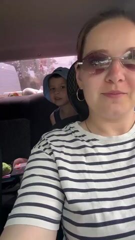 A woman wearing sunglasses and a striped shirt is in the front seat of a car, holding a phone to take a selfie. A young boy wearing a blue hat is in the back seat behind her, peeking over her shoulder and giving a thumbs-up. The car appears to be in motion, as indicated by the blurry background. The interior of the car is visible, including the seats and the car's headrest. The overall scene suggests a casual outing or road trip.