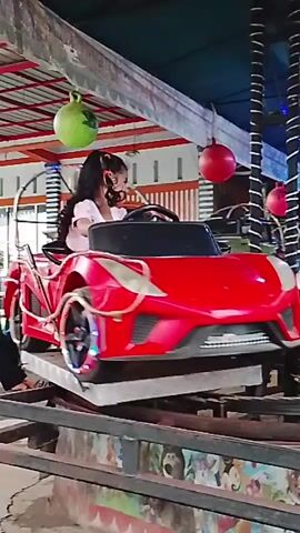 This video shows a slow-moving carnival ride designed for children. A girl with long, dark, curly hair, wearing a light pink shirt, sits in the driver's seat of a bright red toy sports car. The car is positioned on a track that appears to be a circular platform that rotates, causing the cars to move in a circle. In front of the red car, a green toy jeep is visible on the track, moving in the same direction. Both vehicles are moving slowly, and the ride appears to be indoors or under a covered structure, as evidenced by the overhead lighting and supports. The background includes various decorations, such as red and green spheres hanging from the ceiling and a structure with twinkling lights. The ride is designed to simulate driving, with the children in the vehicles appearing to be steering. The overall scene depicts a typical amusement park attraction for young children.