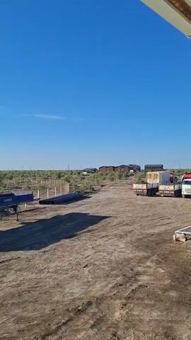 The video shows an outdoor scene with a bright blue sky and a dusty, unpaved ground. In the foreground, there is a long, blue flatbed trailer with three axles. Next to the trailer, there are several long, dark cylindrical objects, likely pipes, lying on the ground. In the middle ground, there are some buildings in the distance, a white truck with a box trailer, and another smaller white truck. The background is a flat landscape with a fence and sparse vegetation. The overall impression is of a work or storage area, possibly in a rural or industrial setting.