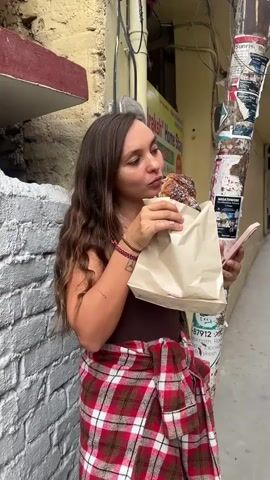 A woman with wavy brown hair is standing on a street corner eating something out of a tan paper bag. She’s holding a cell phone in one hand and the bag in the other, as she takes a large bite of the food. The woman is wearing a brown tank top and a red and white plaid wrap around her waist. She is leaning against a wall made of white bricks, and behind her is a yellow wall with a sign reading “Draksh Home Stay.” There is a utility pole to her right covered in posters and flyers. The street appears to be a narrow alleyway or sidewalk.