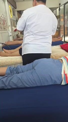 A man in a white uniform stands over a person lying on a massage table. The person on the table is wearing blue pants and a striped shirt, and is lying face down. The room appears to be a spa or massage parlor, with several other massage tables visible in the background, some of which are occupied by other individuals. There are chairs and trash bins in the background, as well as windows that let in natural light. The overall impression is that of a professional service setting.