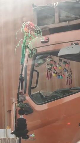 The video shows a close-up view of the front passenger side of an orange truck. The truck is decorated with various ornaments. On the exterior, near the top of the windshield, there's a cluster of pink and green decorative strings and a red flower-like ornament. Hanging from the interior of the windshield is a string of colorful light bulbs and small decorative ornaments with tassels. A large black tassel-like ornament hangs from the side mirror attachment. The background is blurry, suggesting movement or a shallow depth of field, and appears to be a vertical, light-colored surface, possibly another vehicle or structure. The lighting suggests it is daytime, with some glare or dust in the air. No people are visible.