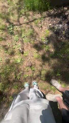 The video opens with an overhead shot showing two pairs of feet standing on dry grass mixed with green patches. Two small, dark, irregular objects are visible on the ground between the feet. Tree shadows dappling the ground suggest a sunny day.

The scene then transitions to a selfie-style shot of a man and a woman standing in a forest. The man, on the left, wears a black hooded sweatshirt with a small red emblem on the chest, dark sunglasses, and has long hair and a beard. He looks directly at the camera. The woman, on the right, wears a light grey hooded sweatshirt, dark sunglasses, and a light green bucket hat. She is looking slightly off-camera to her right. Both subjects are positioned against a backdrop of tall trees with green foliage, indicating a wooded area or forest. The lighting is bright and natural, consistent with a clear, sunny day. No visible text is present.