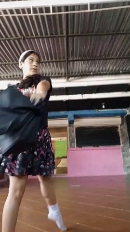 This video shows a young girl dancing in an open-air covered space. She is wearing a black t-shirt, a patterned skirt, and white socks. She is also wearing a pink headband. The floor is tiled, and there is a corrugated metal roof above. In the background, there are pink walls and open doorways with barred windows, suggesting a somewhat rustic or unfinished building. The girl is moving energetically, her skirt swirling around her. The lighting suggests it is daytime. There is no visible text in the video. The context provided indicates the location is El Espinal, Venezuela.