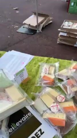 The video shows a cheese stand at an outdoor market. There are several types of cheese on display, including individually packaged cheese wedges, goat cheese logs, and wheels of cheese. A man is working behind the table, possibly preparing food. There are boxes and crates stacked nearby, and a few people can be seen walking around the market in the background.