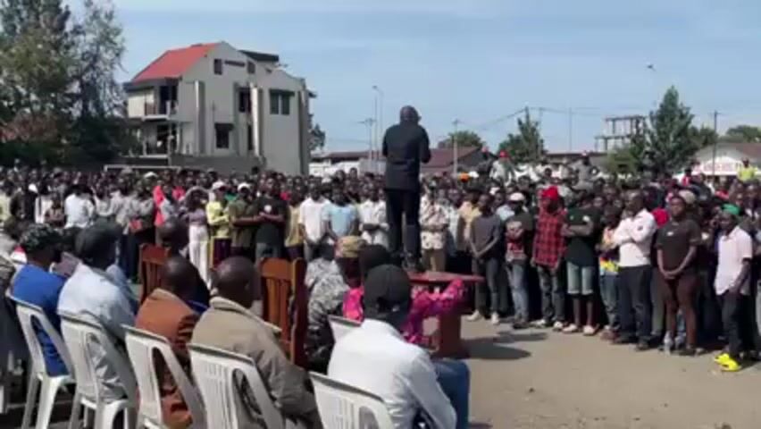 Here is a concise description of the video:

A man in a black suit is speaking to a large crowd outdoors. He is standing on a small table, addressing the crowd with a microphone in his hand. The crowd consists of many people, mostly men, standing and listening attentively. In the foreground, a row of people are seated in white plastic chairs. The setting appears to be an urban area, with buildings and trees visible in the background. The sky is blue with some clouds.