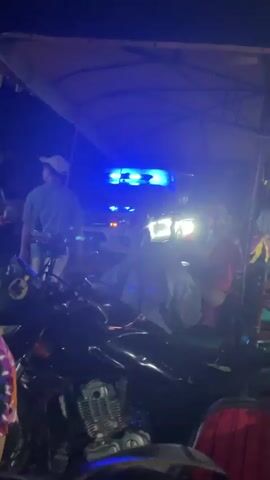 At night, the scene is illuminated by the flashing blue and orange lights of an emergency vehicle visible behind a gathering of people and motorcycles under a shelter. A man in a light-colored shirt walks toward the emergency vehicle. The area seems crowded, with several people standing or sitting around the motorcycles. The shelter has a light-colored roof, and the ground appears dark.