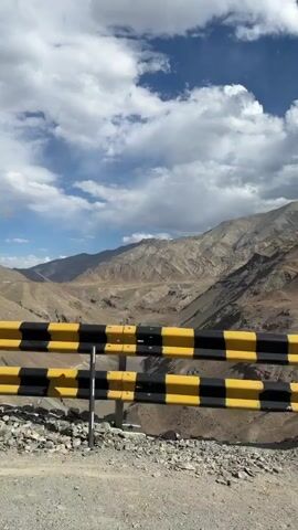 The video shows a drive through a mountainous region. The camera is mounted on a vehicle, likely a motorcycle, as suggested by the handlebar visible in the foreground. The road is paved and winds through steep, rocky hillsides. Guardrails, some yellow and black striped and others white with red stripes, line the edges of the road. A road sign warning of falling rocks is visible on the left side of the road. The sky is blue with scattered white clouds. The terrain appears arid and sparsely vegetated. The video pans and moves forward, giving the impression of a journey.