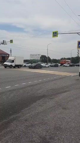 A traffic intersection with multiple lanes and crosswalks is depicted under a cloudy sky. Several cars are present, including a white truck on the left, a dark sedan in the center crossing the intersection, and other cars, including a white one, moving in various directions. Pedestrian crossing signs are visible, with one on the right showing a blue and white pedestrian symbol. A traffic light is also visible on the left. In the background, a large sign with text and images is mounted on a green fence, and a red truck is stopped in the distance. The road surface is asphalt with white lane markings and a yellow and white striped crosswalk.