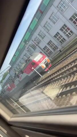 The video shows a train moving along a railway track, seen through a window. The train is painted in a combination of red, white, and grey, with the letters "PЖД" visible on its side. The train tracks run parallel to a large, light grey building with many windows. The sky is blue and clear, and the scene appears to be well-lit by sunlight. There is a figure walking along the side of the building. The camera remains static, and the focus is on the passing train and the adjacent building. The video seems to have been taken from within another train.