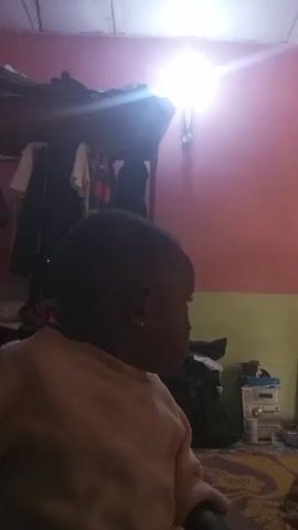 A close-up shot shows a baby with short black hair and a light brown long-sleeved shirt looking towards the right. The baby has a small earring in their left ear. The background features a room with a pink and green wall. A wooden shelf with items and clothes on it is visible above. Below the shelf is a green bag and an electronic device on the floor. The lighting in the room is bright, with a light source above the shelf. The baby is sitting on a patterned mat on the floor.