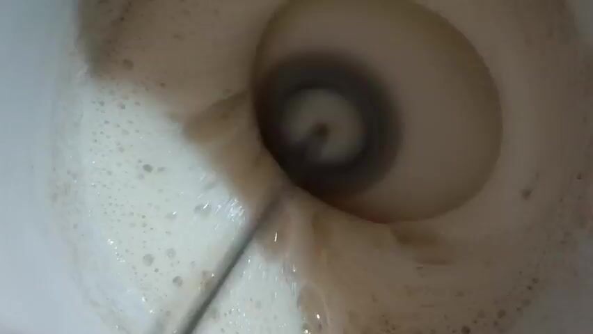 The video shows an overhead view into a light-colored liquid being mixed with a small electric mixer. The liquid is foamy.