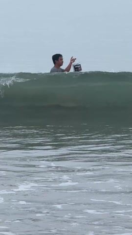 A medium shot captures a man standing in the ocean, holding a can or bucket, possibly of paint, with the brand name "Hidden" visible. He is wearing a grey t-shirt and appears to be gesturing or speaking. Waves crash around him, with a misty, overcast sky in the background. A boat is vaguely visible in the distance.