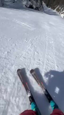 The video shows a first-person perspective of someone skiing downhill on a sunny day. The skier is wearing pink and teal pants, and the skis are visible in the foreground. The ski slope is covered in snow and surrounded by trees on both sides. In the distance, mountains can be seen with patches of snow on them. The sky is blue with some clouds. The skier's shadow is visible on the snow, moving along with them as they ski downhill.