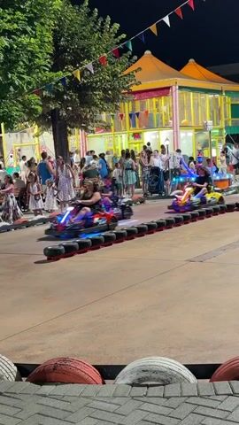 The video shows a night scene at an amusement park or outdoor entertainment venue. In the foreground, a go-kart track is visible, with several go-karts racing. Some of the go-karts have illuminated lights. In the middle ground, there are many people standing around watching the race or waiting in line. Behind the crowd, there are colorful amusement rides and structures. The environment is outdoors, with trees and string lights visible. The overall atmosphere appears to be lively and festive.
