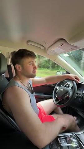 A man with short brown hair and stubble is sitting in the driver's seat of a car, looking at the camera and smiling. He is wearing a grey tank top and pink shorts, and a silver chain around his neck. His right hand is on the steering wheel, and his left hand is resting on his knee. The car is moving, as indicated by the blurred greenery outside the window and the movement of the steering wheel. The dashboard of the car is visible, with a large touchscreen display. The video cuts to a view from the dashboard, showing the road ahead. The car is on a paved road surrounded by trees and fields. The sky is blue with some clouds. Another car is visible in the distance on the same road.