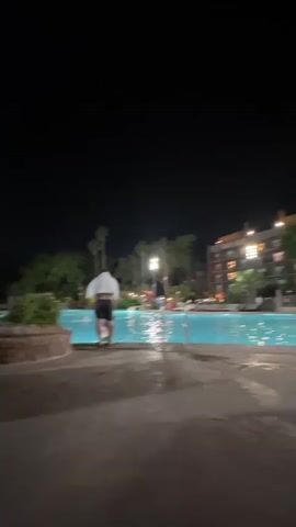 The video shows an outdoor swimming pool area at night. The pool is lit up and appears to be empty. A person walks away from the pool in the beginning of the video. There are lounge chairs with cushions near the camera. There are umbrellas, palm trees, bushes, and buildings visible in the background. The overall lighting is low, and the sky is dark.