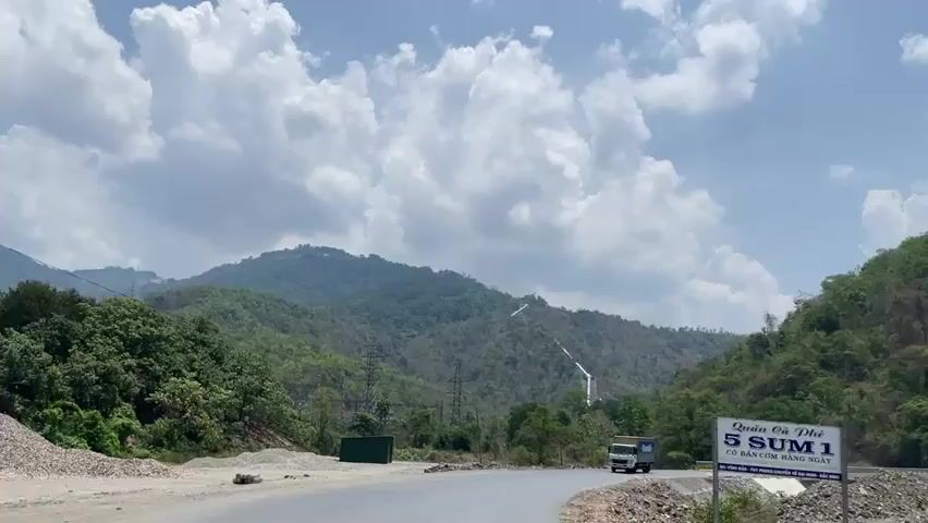 The video shows a road in a mountainous area. A small, white truck with "Vinamilk" written on its side drives along the road and passes the camera. The road has a bridge and there are mountains covered in trees on either side of the road. There are power lines visible. The sky is blue with white clouds. There is a sign at the side of the road with Vietnamese writing. The road appears to be relatively empty.
