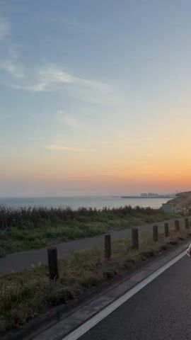 The video captures a scenic coastal view during sunset, likely from a moving vehicle. The scene features the ocean on the left, with a calm sea reflecting the warm hues of the sky. A road runs along the right side of the frame, bordered by dry grass and vegetation. A pathway, possibly for cyclists or pedestrians, is visible between the road and the grassy embankment.

Several objects are prominent: a tall lamppost with a blue bicycle sign is visible on the right, and a cellular tower stands further down the road. A circular speed limit sign displaying "50" is also clearly visible. In the distance, a coastline with some buildings and structures can be made out. The sky is a gradient of soft blues and oranges, indicative of a sunset. The weather appears clear and pleasant.

The activity taking place is travel, as the perspective suggests motion. A side mirror of the vehicle shows a reflection of a person, hinting at the presence of occupants. The overall mood is serene and tranquil, enhanced by the beautiful sunset colors and the peaceful coastal setting. The location context of Ovingdean, United Kingdom, suggests a coastal drive along the English Channel.