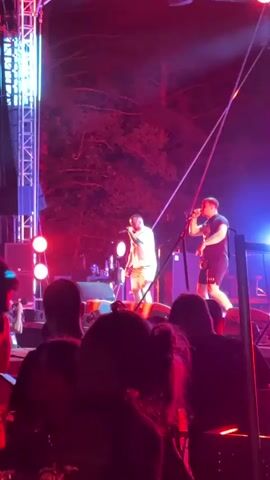 A concert is taking place outdoors at night. Two male performers are on a stage, each holding a microphone and singing. One performer, wearing a white t-shirt and shorts, stands center-left. The other, wearing a black t-shirt and shorts, stands center-right. Both are illuminated by stage lights that cast red and blue hues on the scene. Behind them, trees are visible, silhouetted against the bright lights. In front of the stage, a crowd of people can be seen, their backs to the camera, watching the performance. The overall atmosphere appears energetic and lively, with the audience engaged in the concert.