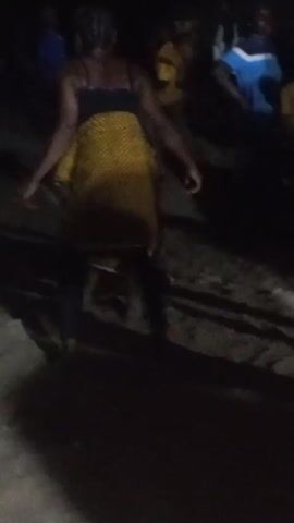 The video shows a person in the center wearing a black top, black pants, and a patterned yellow and brown wrap. They are dancing, with their arms extended to the sides. Another person wearing a black top and a red and yellow knit hat enters the frame on the right and briefly interacts with the dancer. The scene appears to be at night outdoors on what looks like a sandy or dirt surface, with other blurry figures in the background. The lighting is dim and possibly from a distant source.