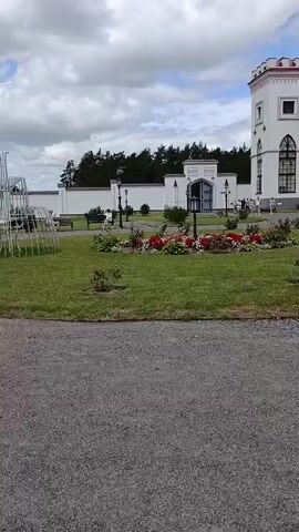The video shows a park-like setting with a gravel path in the foreground. In the mid-ground, there is a grassy area with a decorative white fountain structure. Several people are present in the mid-ground, some sitting on benches and others walking or playing. A flower bed with red flowers is visible to the right of the fountain. In the background, there is a white wall with a gate and trees beyond that. The sky is cloudy. The camera pans slightly to the left and then to the right. The actions observed include people sitting, walking, and interacting with each other.