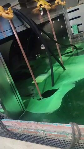 The video shows a close-up, slightly shaky view of a vat filled with bright green liquid, likely some form of chemical solution. The liquid has an opaque, viscous appearance, with some areas appearing darker and more concentrated. Several black, flexible tubes are submerged in the green liquid, with their ends disappearing beneath the surface. These tubes are supported by copper-colored rods that extend vertically from the liquid. Above the vat, metal structures with yellow lifting hooks are visible, suggesting that the vat or its contents are being moved or processed. The overall setting appears to be an industrial environment, possibly a factory or laboratory, given the metallic machinery and the nature of the liquid. There is no clear indication of the time of day or weather conditions. The scene is dominated by the intense green color of the liquid and the metallic elements of the machinery. No people are visible in the frame, and no text is legible. The audio consists of speech in German, with the first few seconds containing the words "Wir sind täglich live" which translates to "We are live daily."
