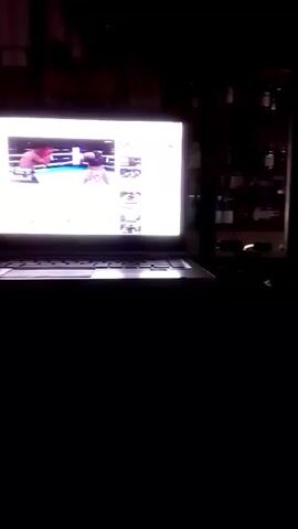 The video begins with a view of a laptop screen, the image indistinct but appearing to show some kind of sports event. The camera then pans to the right, revealing a dimly lit room with a shelf unit containing various objects. There is a light source on or behind the shelf, though the details are obscured by darkness.  The camera then pans downwards towards what appears to be a piece of furniture, such as a couch or chair. The remainder of the video consists of dark, shaky footage. The general environment seems to be inside a cluttered space, with visibility limited due to poor lighting.