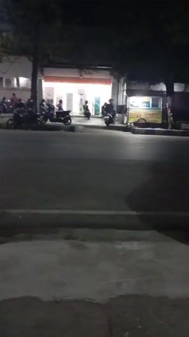 The video shows a street scene at night. A row of shops are visible in the background, illuminated by interior lighting. Several motorcycles are parked in front of the shops and on the side of the road. Some people are standing and walking in front of the shops. Two motorcycles are seen driving past the camera. One motorcycle has its brake lights illuminated. A small food cart is visible on the right side of the street. The road surface is paved, and there is a concrete sidewalk in the foreground. The overall impression is a moderately busy street in a commercial area at night.